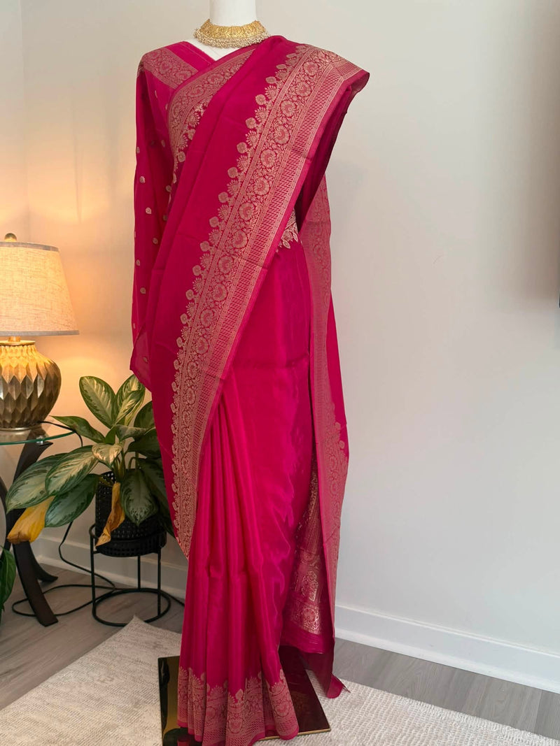 A bright pink silk saree with intricate gold zari embroidery and border detailing, displayed on a mannequin in an indoor setting with a lamp and plant in the background.
