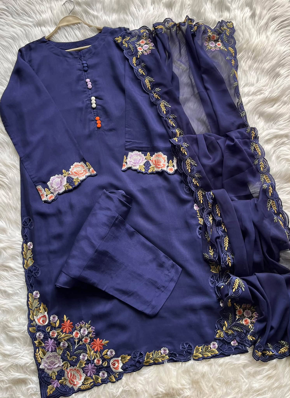A navy blue traditional outfit set with floral embroidery and lace trim, laid flat on a white faux fur background. The set includes a long kurta with embroidered cuffs and hem, matching pants, and a sheer dupatta with lace borders.