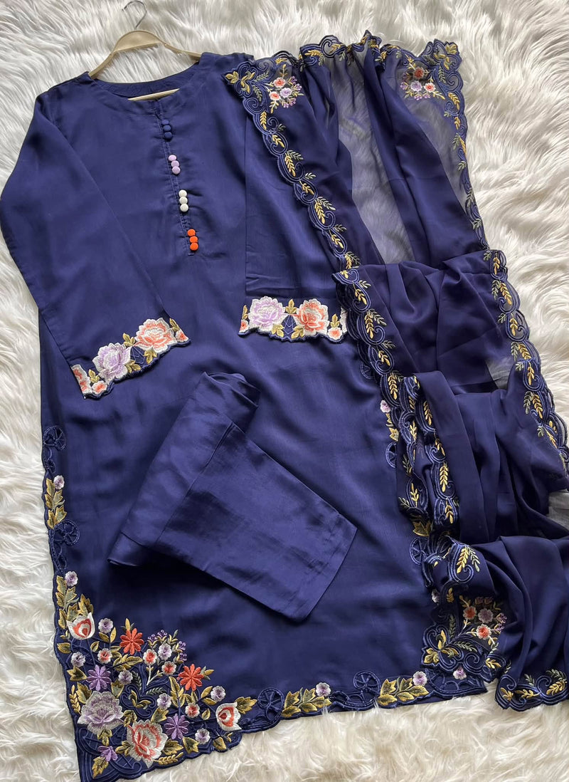 A navy blue traditional outfit set with floral embroidery and lace trim, laid flat on a white faux fur background. The set includes a long kurta with embroidered cuffs and hem, matching pants, and a sheer dupatta with lace borders.