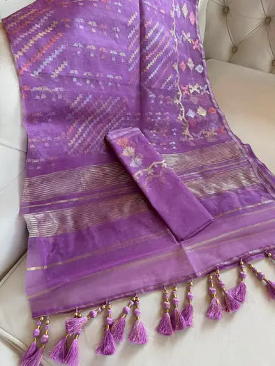 A purple silk saree with intricate gold and multicolor woven patterns, displayed on a cream tufted sofa. The saree features a matching purple fabric sleeve rolled up and a decorative border with purple tassels.