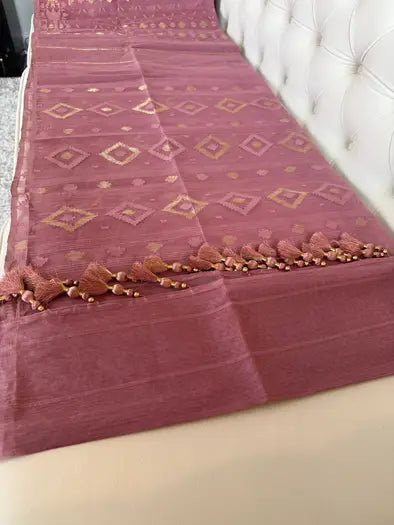 A  rose pink silk saree or fabric runner with gold geometric diamond patterns and a decorative tassel border, draped over a white tufted headboard.