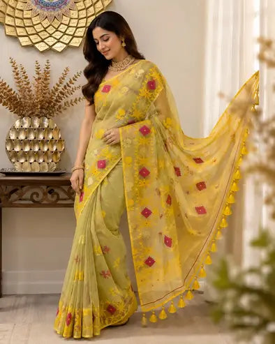 A woman models a yellow and olive green embroidered saree with pink floral accents and a matching yellow border with tassels, set against an indoor background with decorative elements.