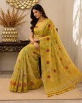 A woman models a yellow saree with red floral motifs and a matching yellow dupatta draped over her lap, set in an indoor styled room with a table and decorative vase in the background.