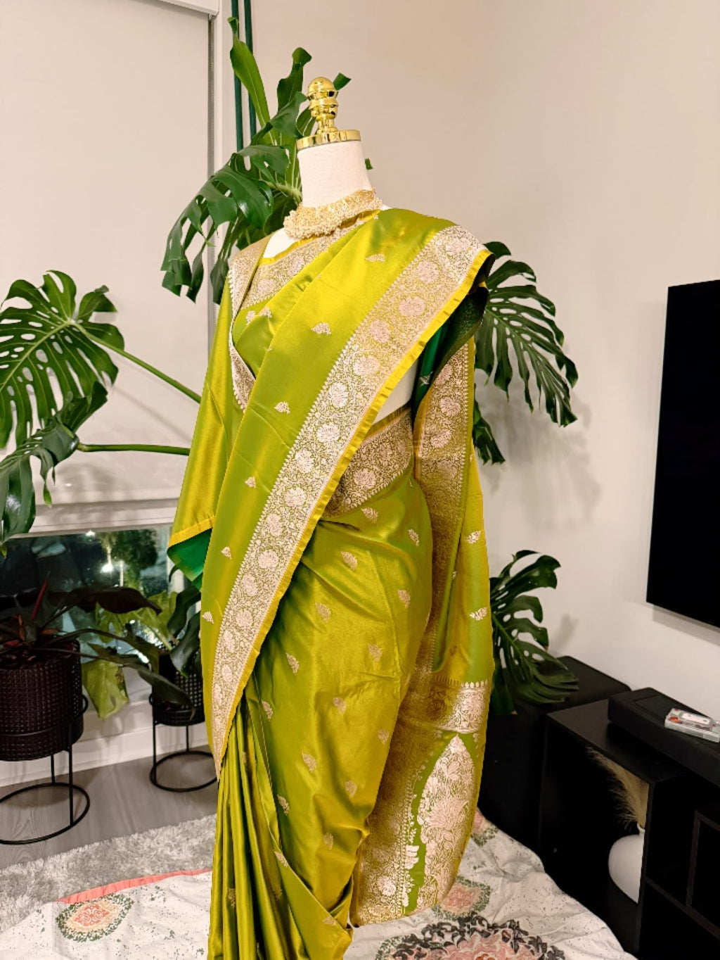 A bright yellow-green silk saree with intricate gold and silver zari embroidery, displayed on a mannequin in a home setting with potted plants and a TV in the background.
