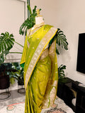 A bright yellow-green silk saree with intricate gold and silver zari embroidery, displayed on a mannequin in a home setting with potted plants and a TV in the background.