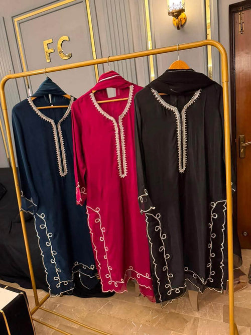 Three long abaya dresses hanging on a gold clothing rack in an indoor showroom. The dresses are shown in navy blue, bright magenta pink, and black, each featuring white embroidered patterns along the neckline and lower skirt.