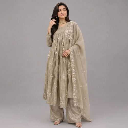 A woman modeling a traditional beige three-piece outfit with matching pants and a sheer embroidered dupatta, photographed against a solid gray studio background.