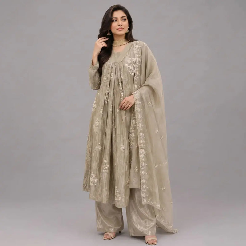 A woman modeling a traditional beige three-piece outfit with matching pants and a sheer embroidered dupatta, photographed against a solid gray studio background.
