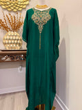 A dark green kaftan dress with ornate gold embroidery around the neckline and a long tassel, displayed on a mannequin in an indoor setting.