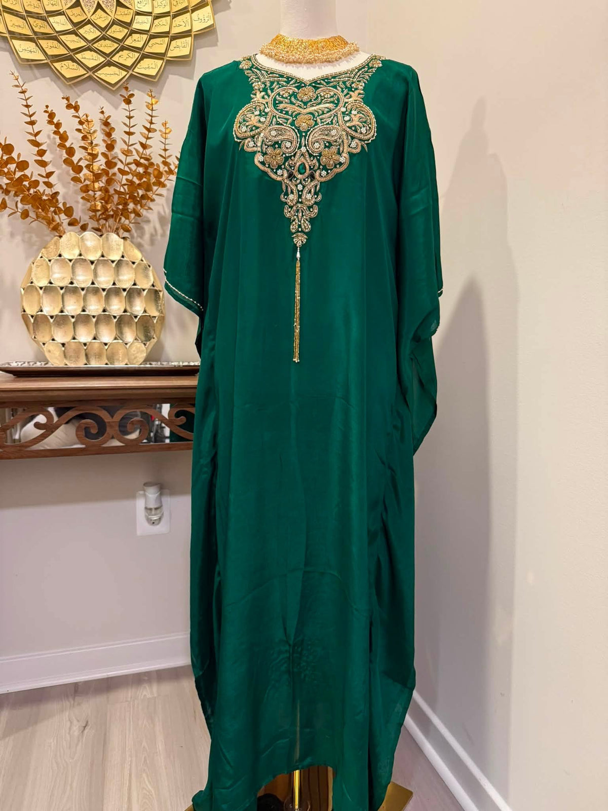 A dark green kaftan dress with ornate gold embroidery around the neckline and a long tassel, displayed on a mannequin in an indoor setting.