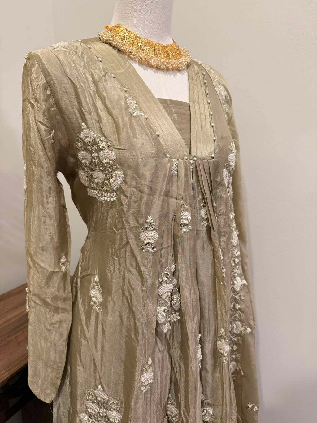 A beige embroidered traditional dress displayed on a mannequin, featuring intricate white floral embroidery and a gold beaded collar, set against a plain light gray wall.