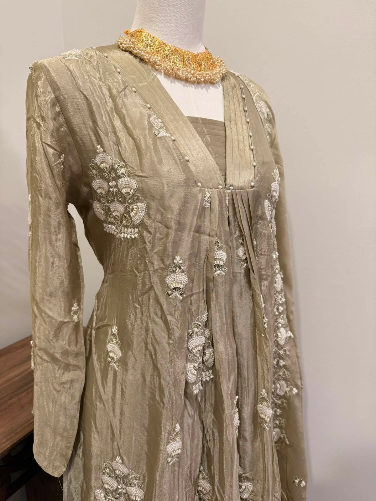 A beige embroidered traditional dress displayed on a mannequin, featuring intricate white floral embroidery and a gold beaded collar, set against a plain light gray wall.
