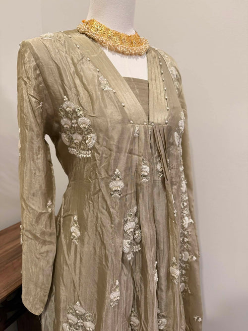 A beige embroidered traditional dress displayed on a mannequin, featuring intricate white floral embroidery and a gold beaded collar, set against a plain light gray wall.