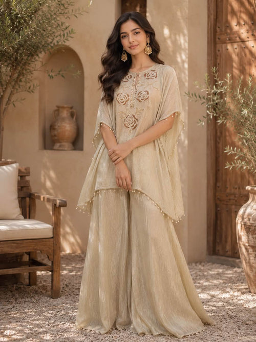 A woman stands outdoors wearing a flowing beige traditional outfit with embroidered floral details on the chest and draped sheer sleeves, paired with wide-leg pants. The setting includes a stucco wall, wooden door, plants, and a chair.