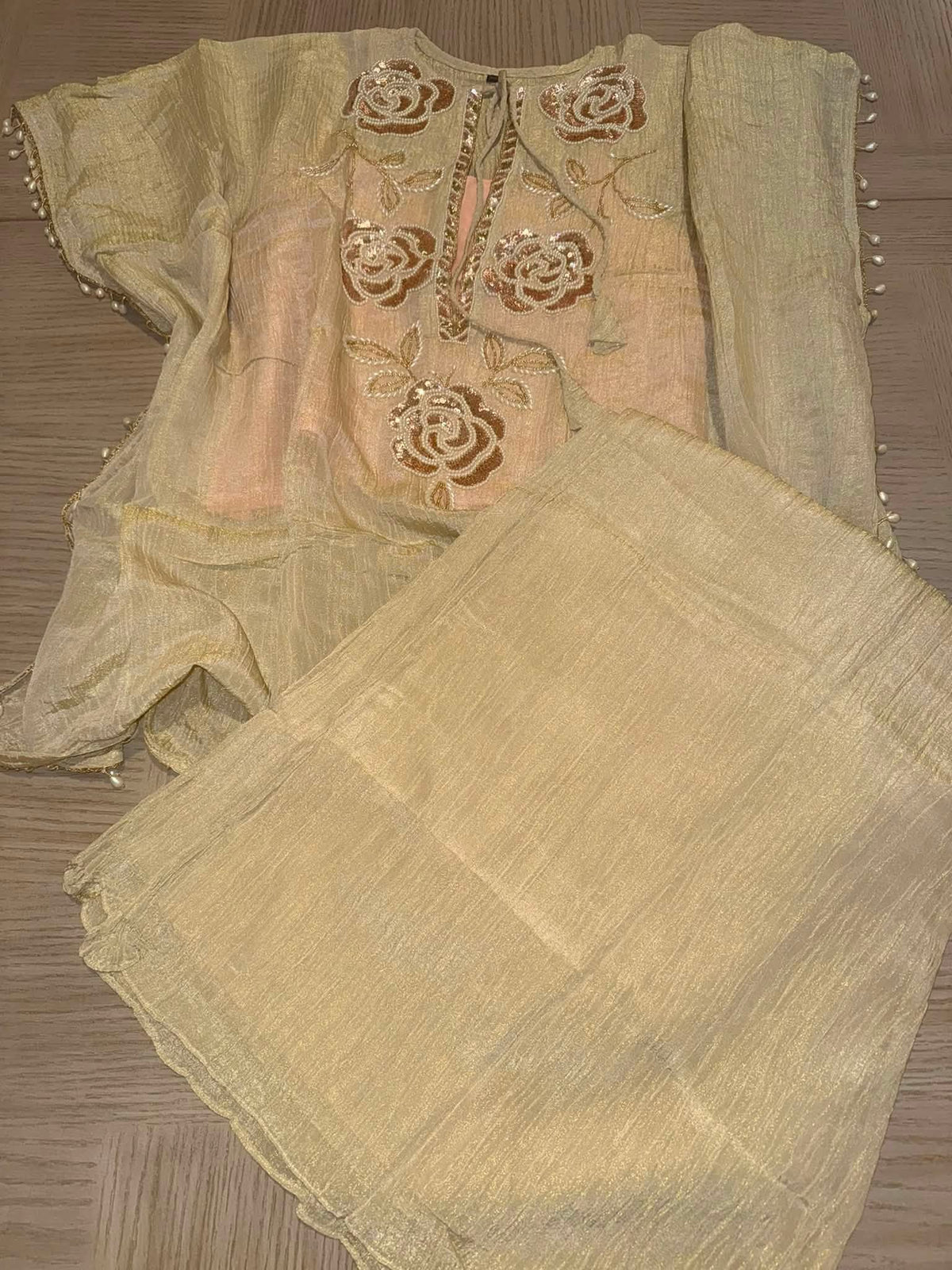 A traditional Indian salwar kameez set in light beige and gold, featuring a sheer embroidered top with rose motifs, a matching dupatta, and textured trousers, laid flat on a wooden floor.