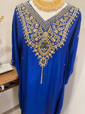 A royal blue kaftan dress with elaborate gold and silver embroidery and green accents around the V-neck, displayed on a mannequin torso with a gold beaded necklace.