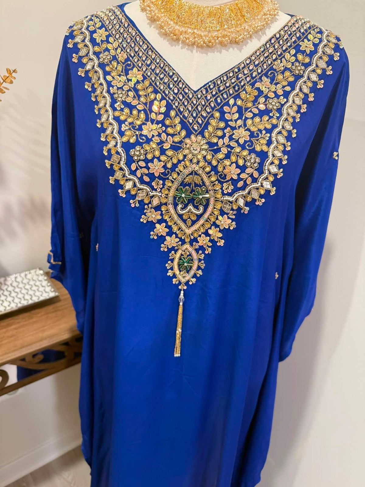 A royal blue kaftan dress with elaborate gold and silver embroidery and green accents around the V-neck, displayed on a mannequin torso with a gold beaded necklace.