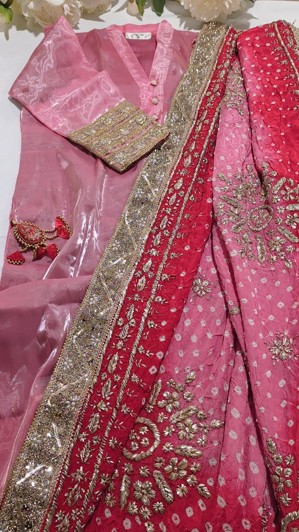 A pink traditional ethnic outfit laid flat, featuring a sheer pink kurta with gold embroidery on the sleeve, a matching pink dupatta with red border and gold motifs, and a red inner layer with gold embellishment.