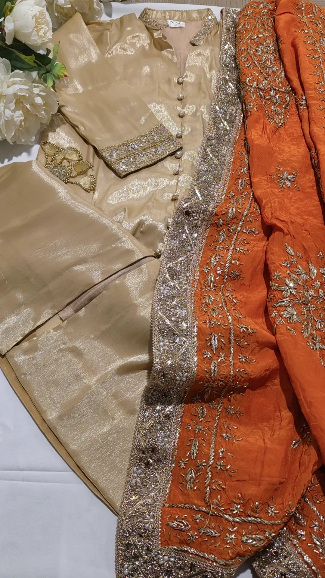 A traditional Indian ethnic outfit laid flat on a white surface, featuring a champagne beige embroidered jacket with matching pants and a bright orange dupatta with ornate gold embroidery and a wide metallic border.