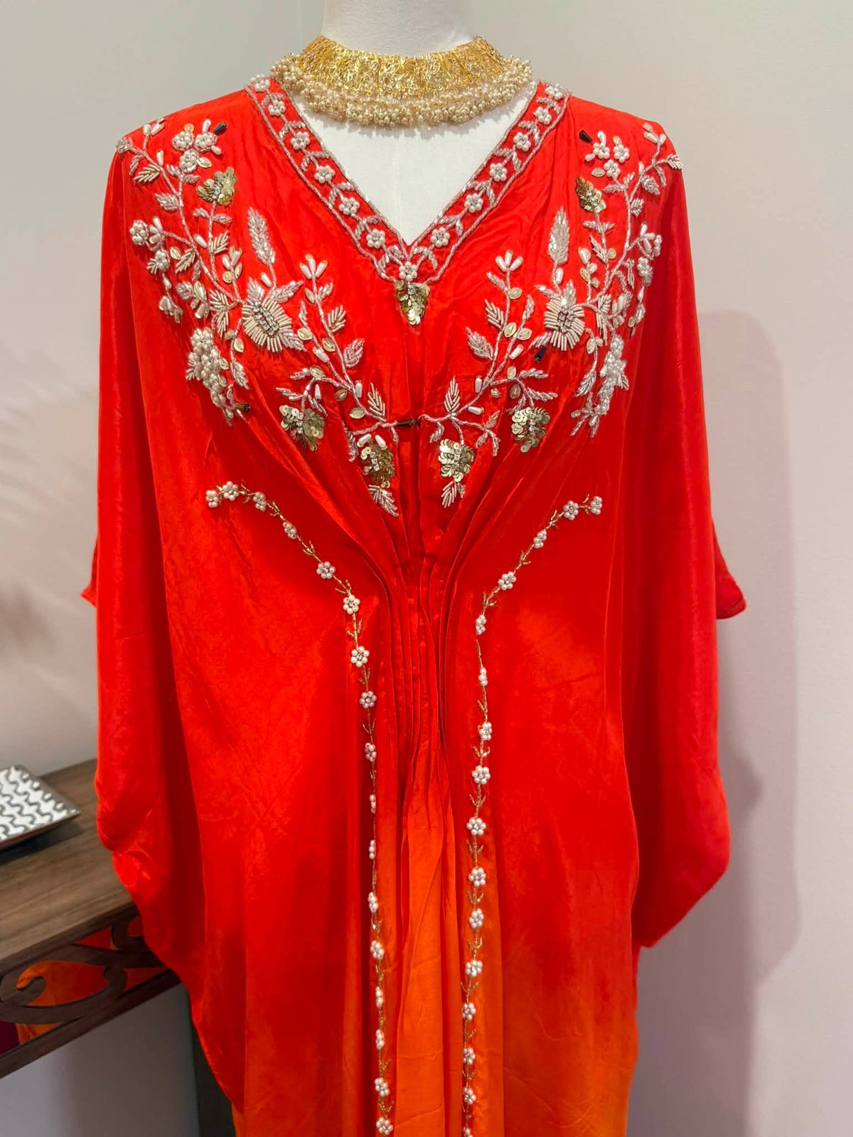 A bright red kaftan dress with intricate silver and green floral embroidery, displayed on a mannequin with a gold beaded collar against a plain light background.