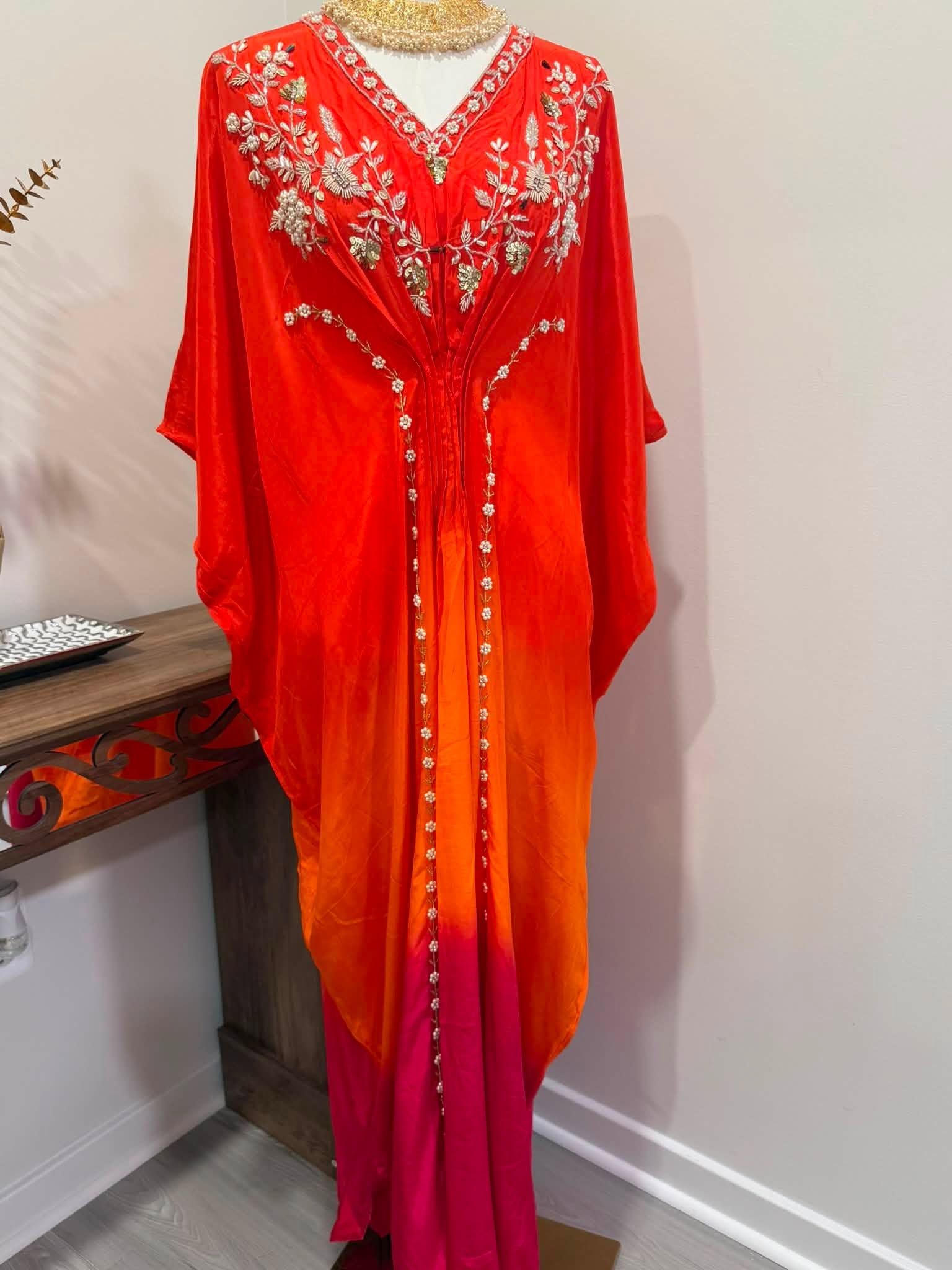 A long red and orange kaftan dress with a magenta hem, featuring silver floral embroidery and bead detailing on the neckline and front center, displayed on a mannequin in an indoor setting.