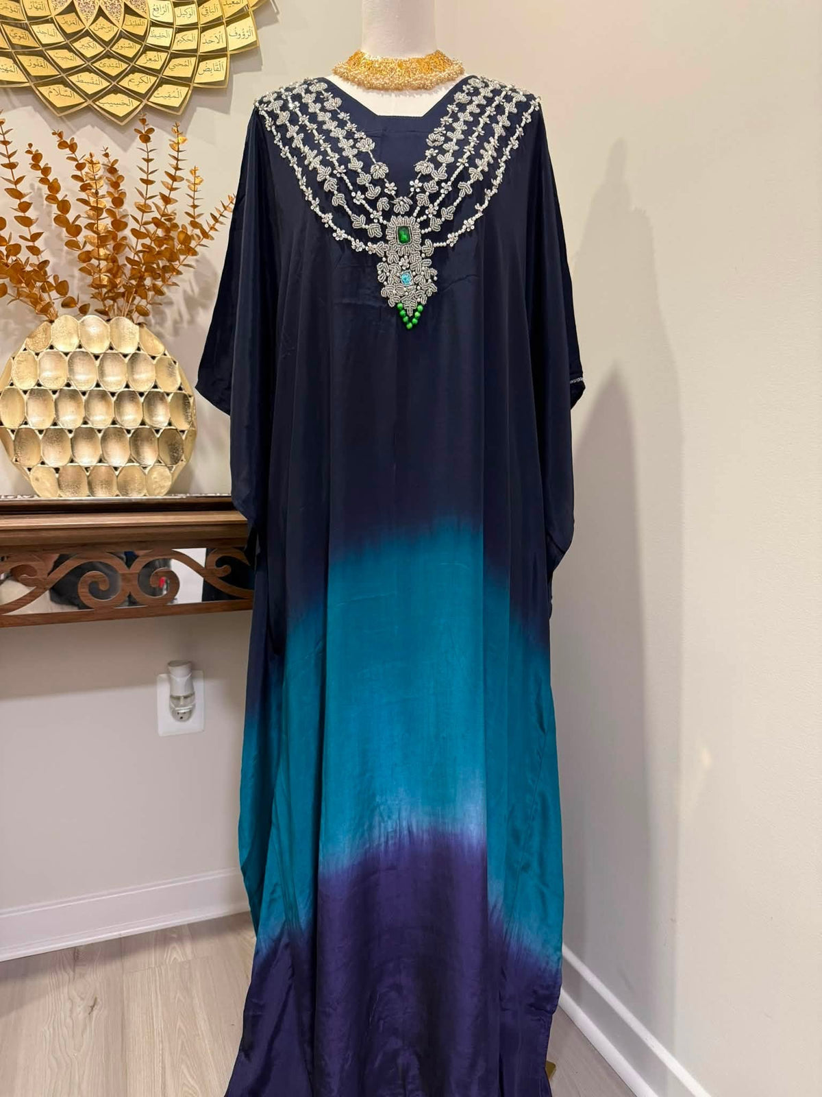 A long flowing caftan or dress displayed on a mannequin indoors, featuring a dark navy upper section that fades into teal and deep purple at the bottom. The neckline is heavily embellished with layered silver beaded chains and a large ornate silver pendant with green and blue stone accents.