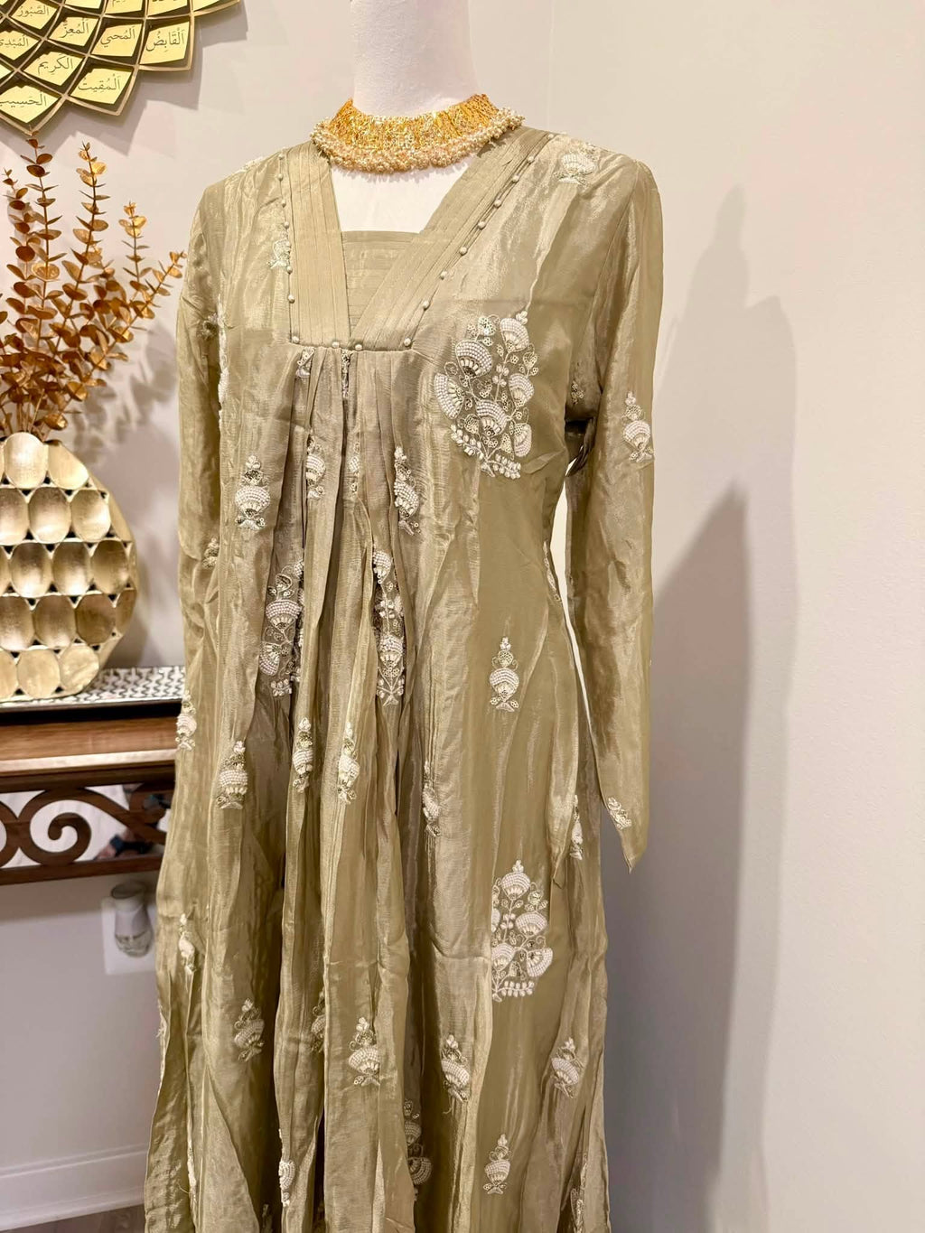 A long-sleeved beige dress with white floral embroidery and a gold beaded collar, displayed on a mannequin in an indoor setting.