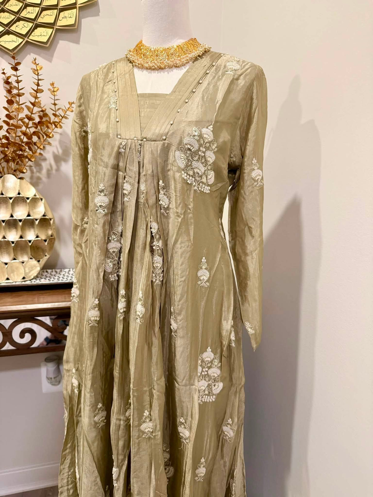 A long-sleeved beige dress with white floral embroidery and a gold beaded collar, displayed on a mannequin in an indoor setting.