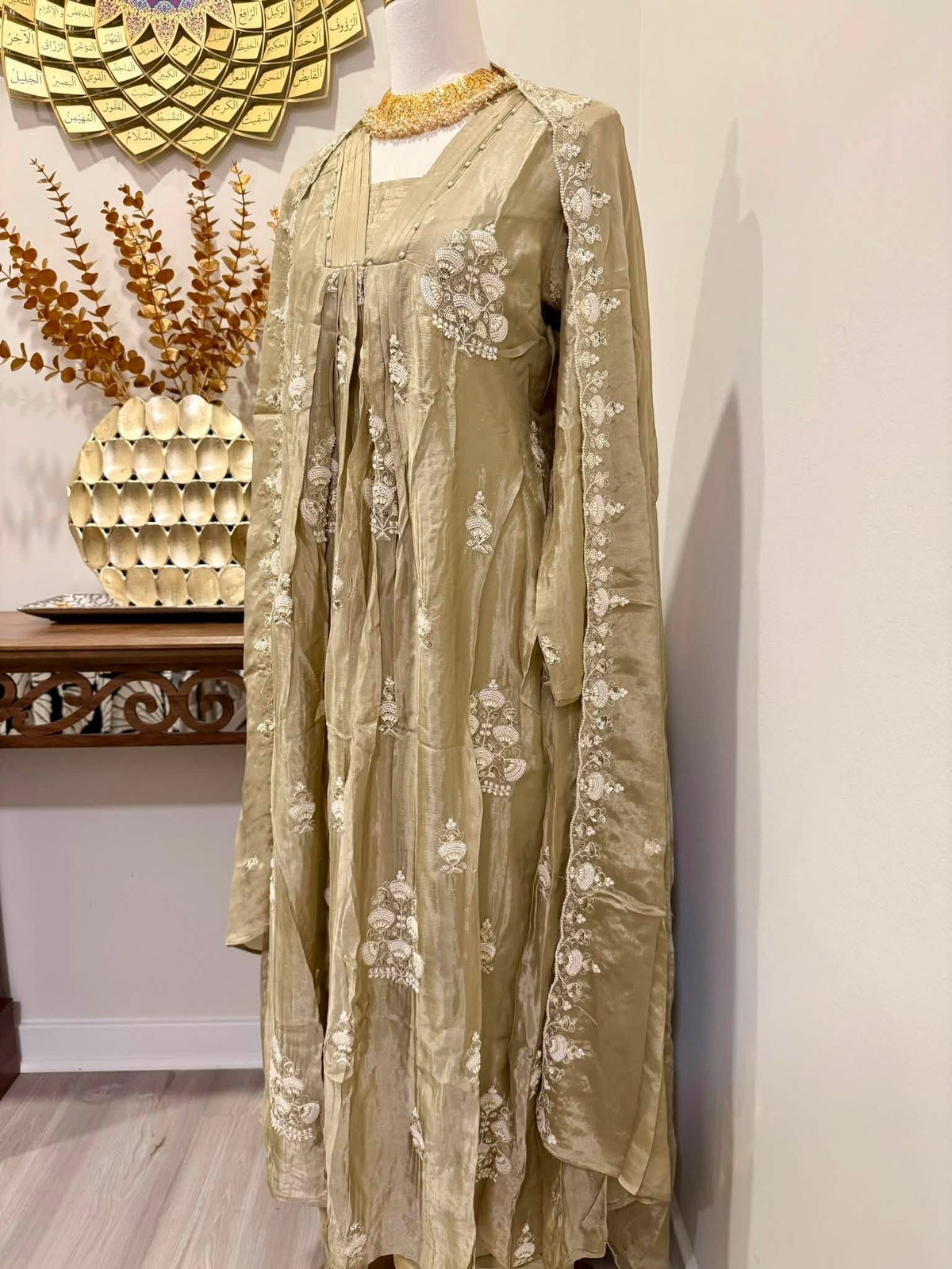 A long-sleeved beige embroidered dress displayed on a mannequin in an indoor setting, featuring intricate white floral embroidery and a gold-toned embellished neckline.