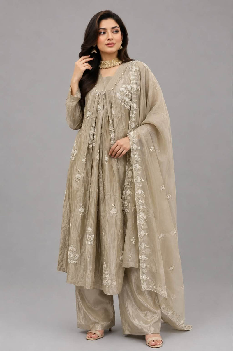 A woman modeling a traditional beige embroidered kurta set with matching wide-leg pants and a sheer dupatta, photographed against a solid gray studio background.
