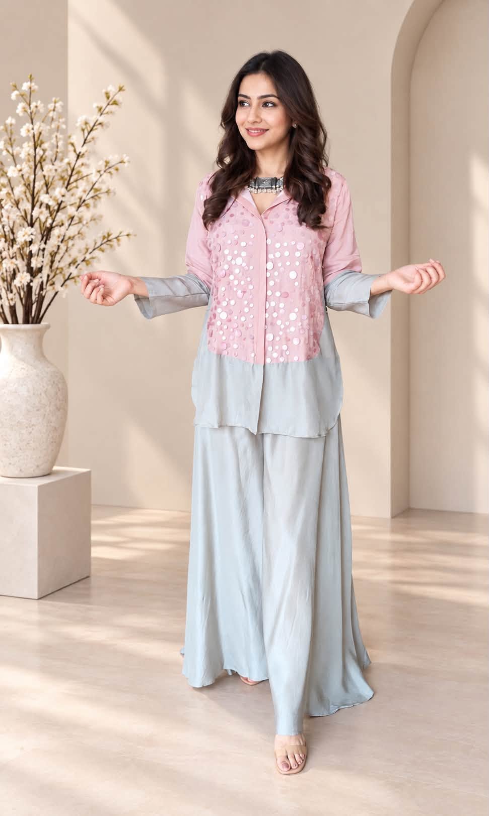 A woman models a two-piece light pink and light blue traditional outfit with a matching long skirt, standing in a warm beige interior setting with a decorative vase of branches on the left.