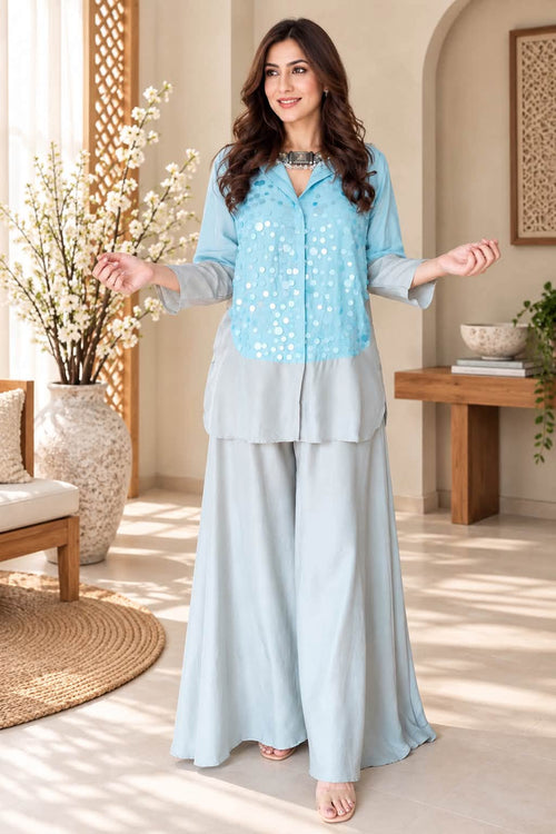 A woman models a light blue two-piece outfit consisting of a long-sleeve button-up top with white printed motifs and matching wide-leg pants, set in a warm, sunlit room with neutral beige walls and wooden furniture.
