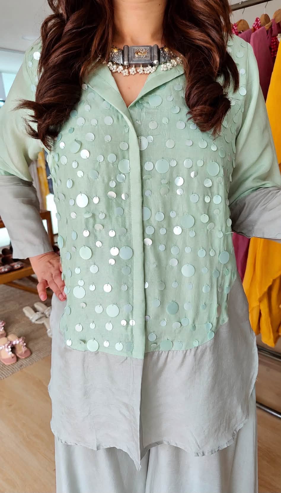 A woman wearing a mint green embellished button-up top with silver mirror work, layered over a light gray tunic and wide-leg pants, in an indoor boutique setting.