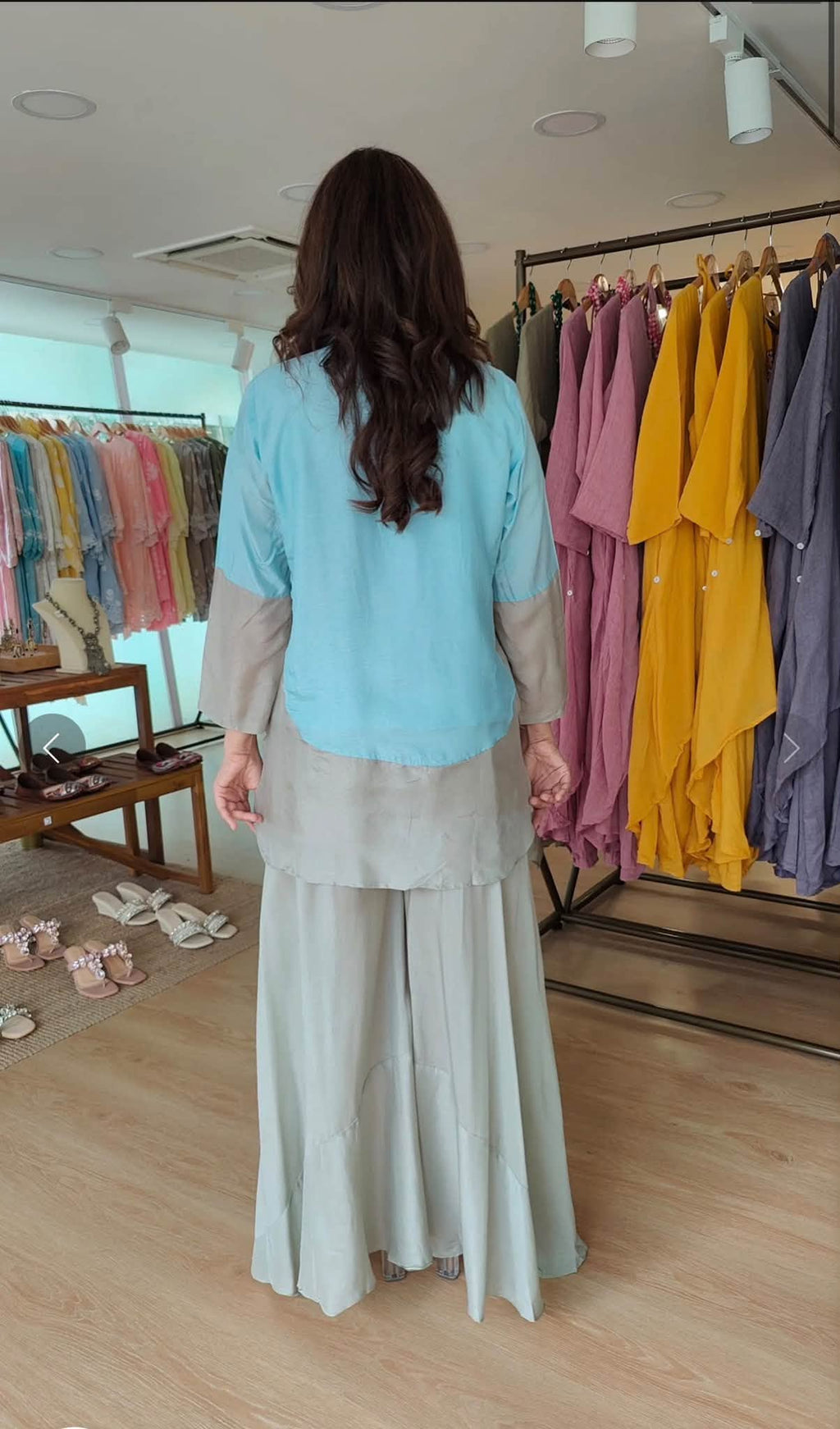 A woman viewed from behind wearing a light blue and beige two-piece tunic with wide-leg light gray-green pants, standing inside a clothing boutique with racks of colorful garments in the background.