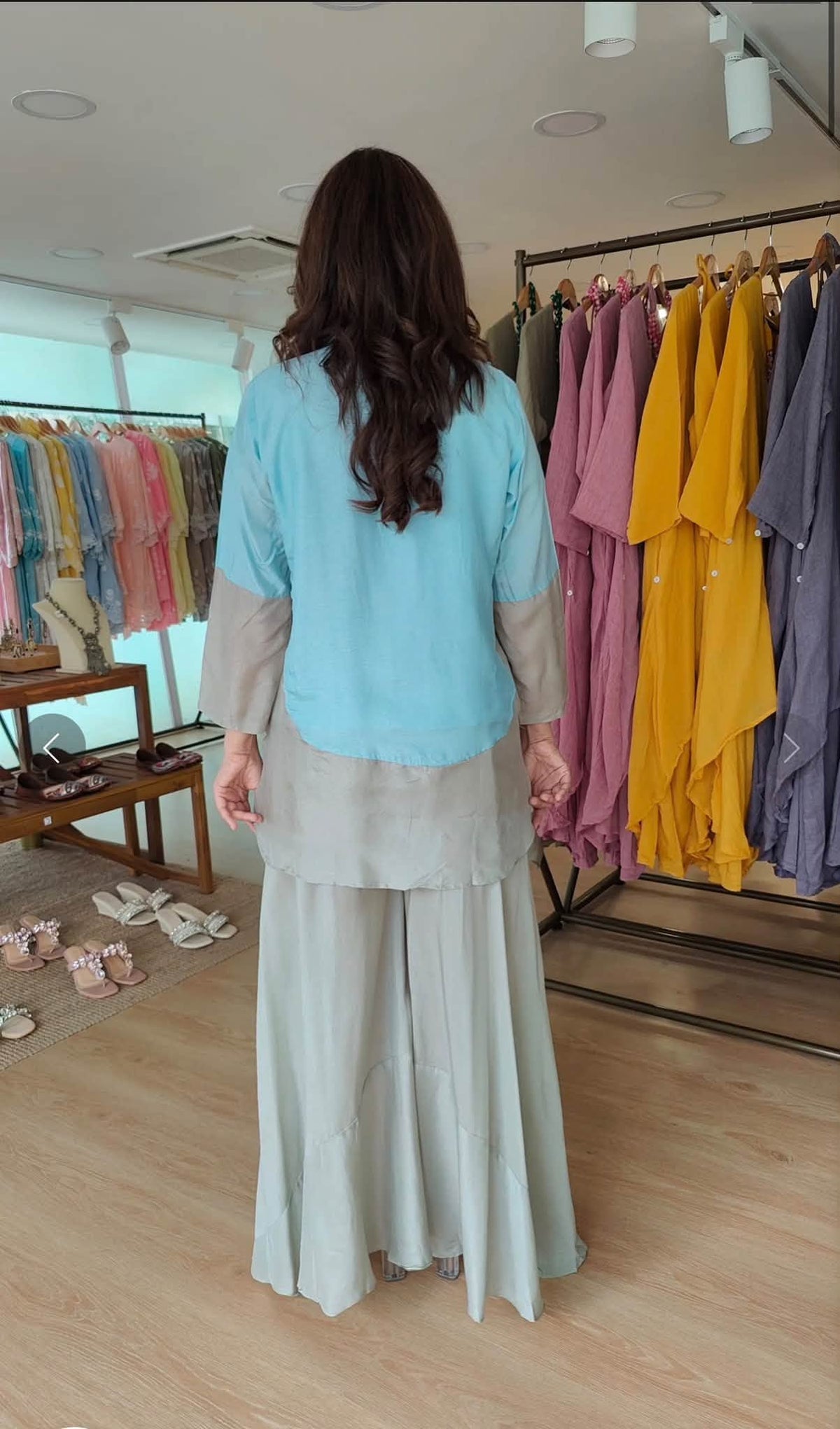 A woman viewed from behind wearing a light blue and beige two-piece tunic with wide-leg light gray-green pants, standing inside a clothing boutique with racks of colorful garments in the background.