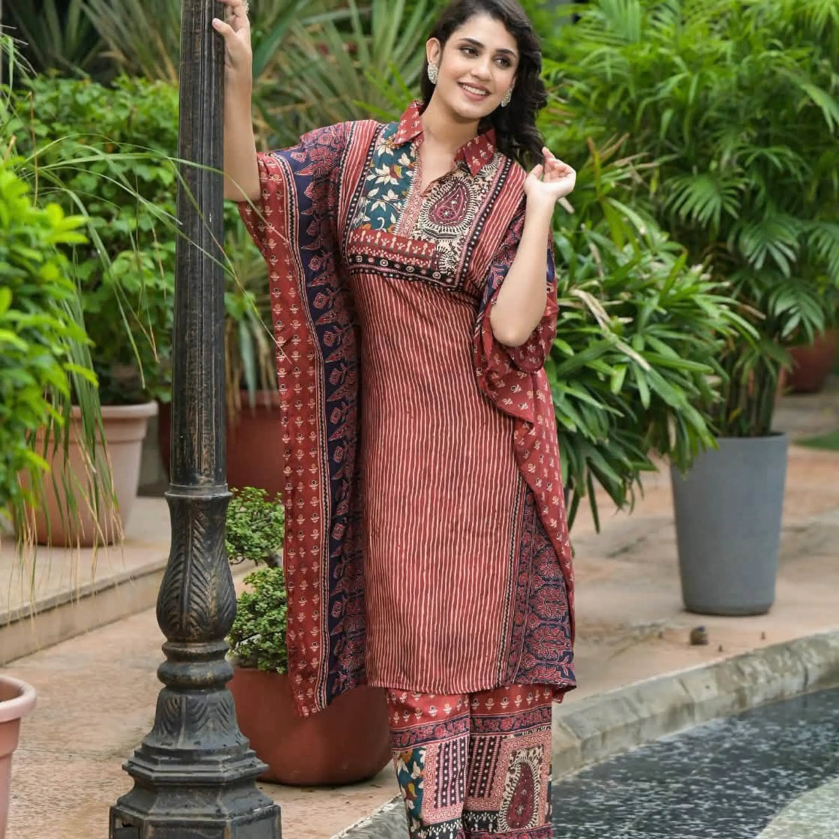 A woman models a rust-red and navy printed ethnic kurta set with matching pants and dupatta, standing next to a black metal lamp post in a garden setting.