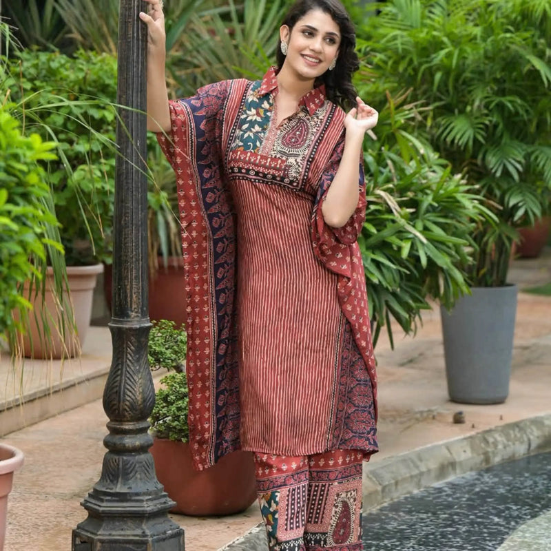 A woman models a rust-red and navy printed ethnic kurta set with matching pants and dupatta, standing next to a black metal lamp post in a garden setting.