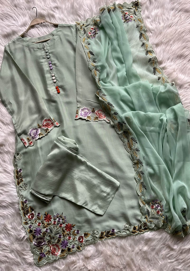A sage green three-piece salwar kameez set with floral embroidery, laid flat on a white faux fur background. The set includes a long kurta with a decorative bead placket, matching trousers, and a dupatta with embroidered borders.