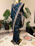 A dark teal blue saree with intricate gold and silver floral patterns, draped on a mannequin in an indoor setting with potted plants and a patterned rug.