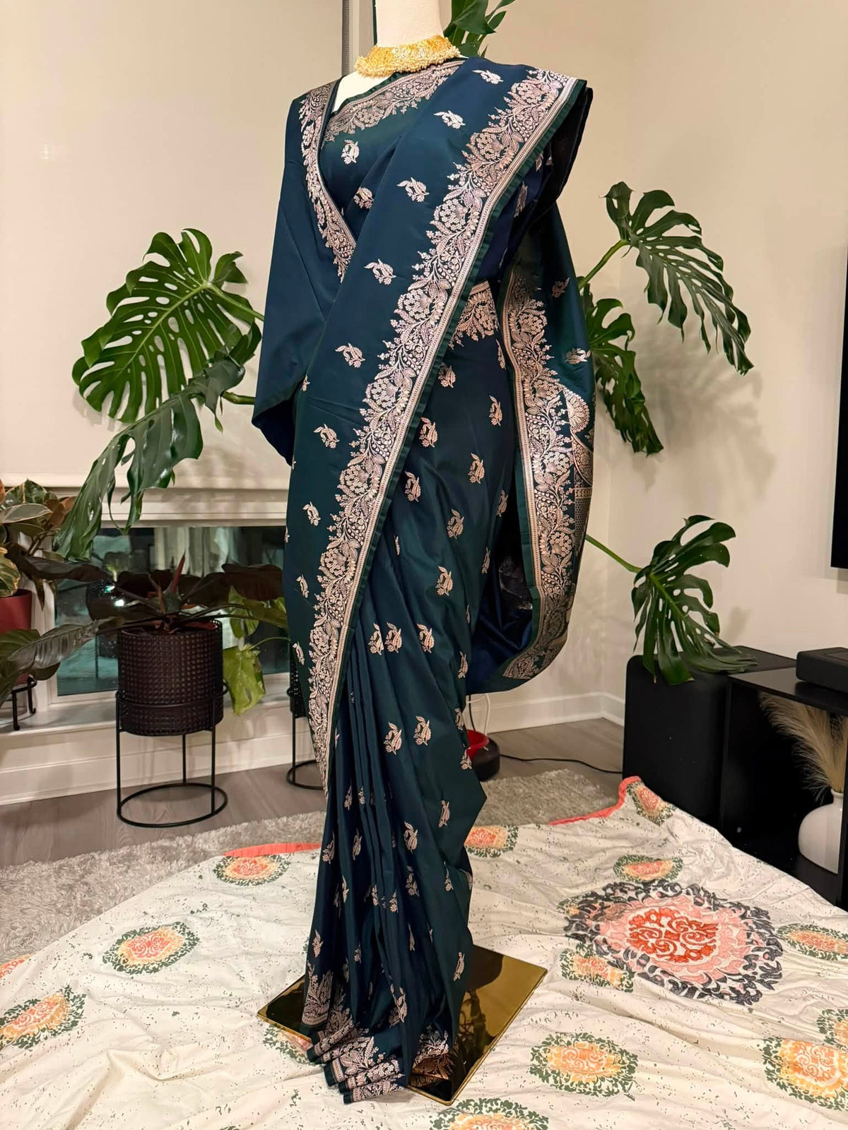 A dark teal blue saree with intricate gold and silver floral patterns, draped on a mannequin in an indoor setting with potted plants and a patterned rug.