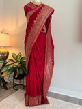A deep red silk saree with intricate gold zari patterns, displayed on a mannequin in an indoor setting with a lamp and plant in the background.