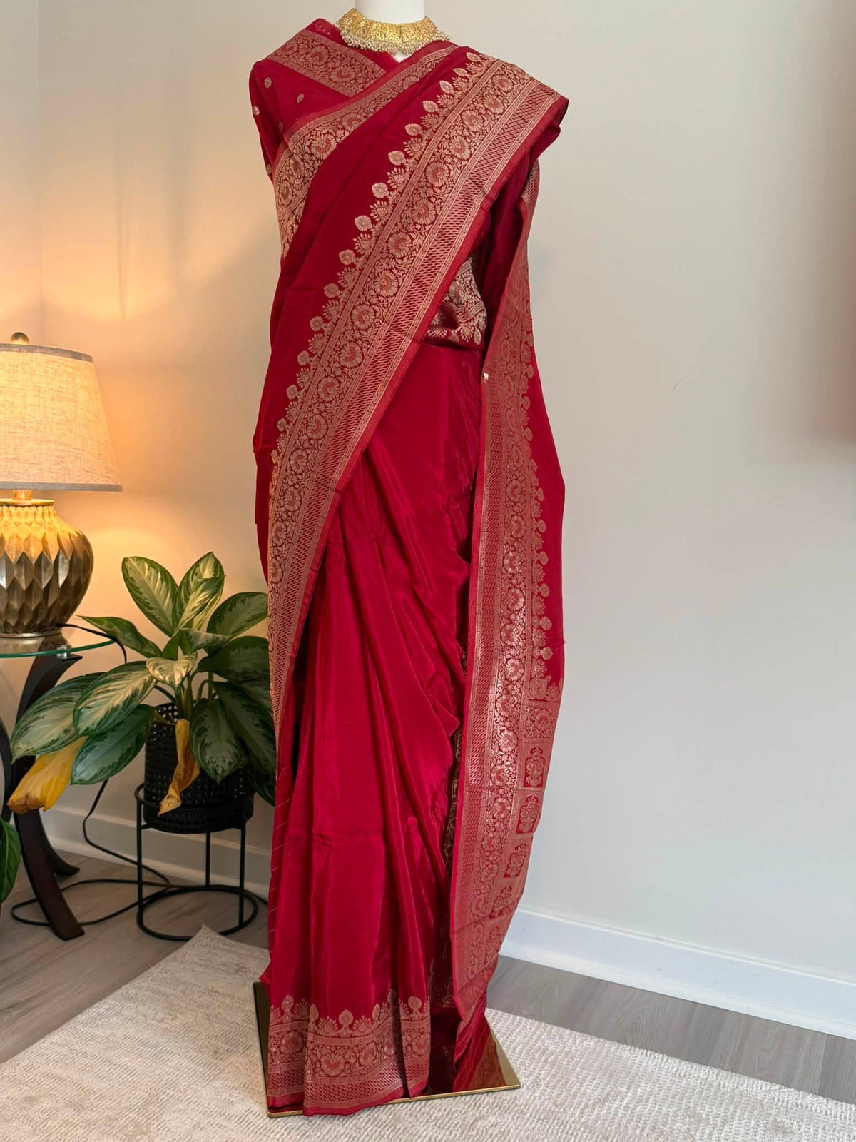 A deep red silk saree with intricate gold zari patterns, displayed on a mannequin in an indoor setting with a lamp and plant in the background.